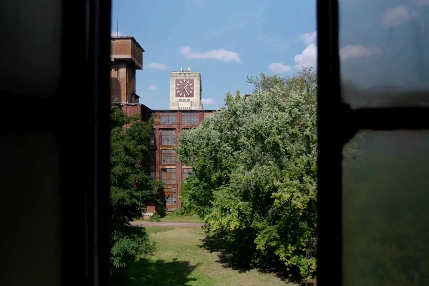 Ein Blick durch ein offenes Fenster auf einen roten Backsteinturm mit einer großen Uhr und blauen Himmel im Hintergrund, umgeben von grünen Bäumen.