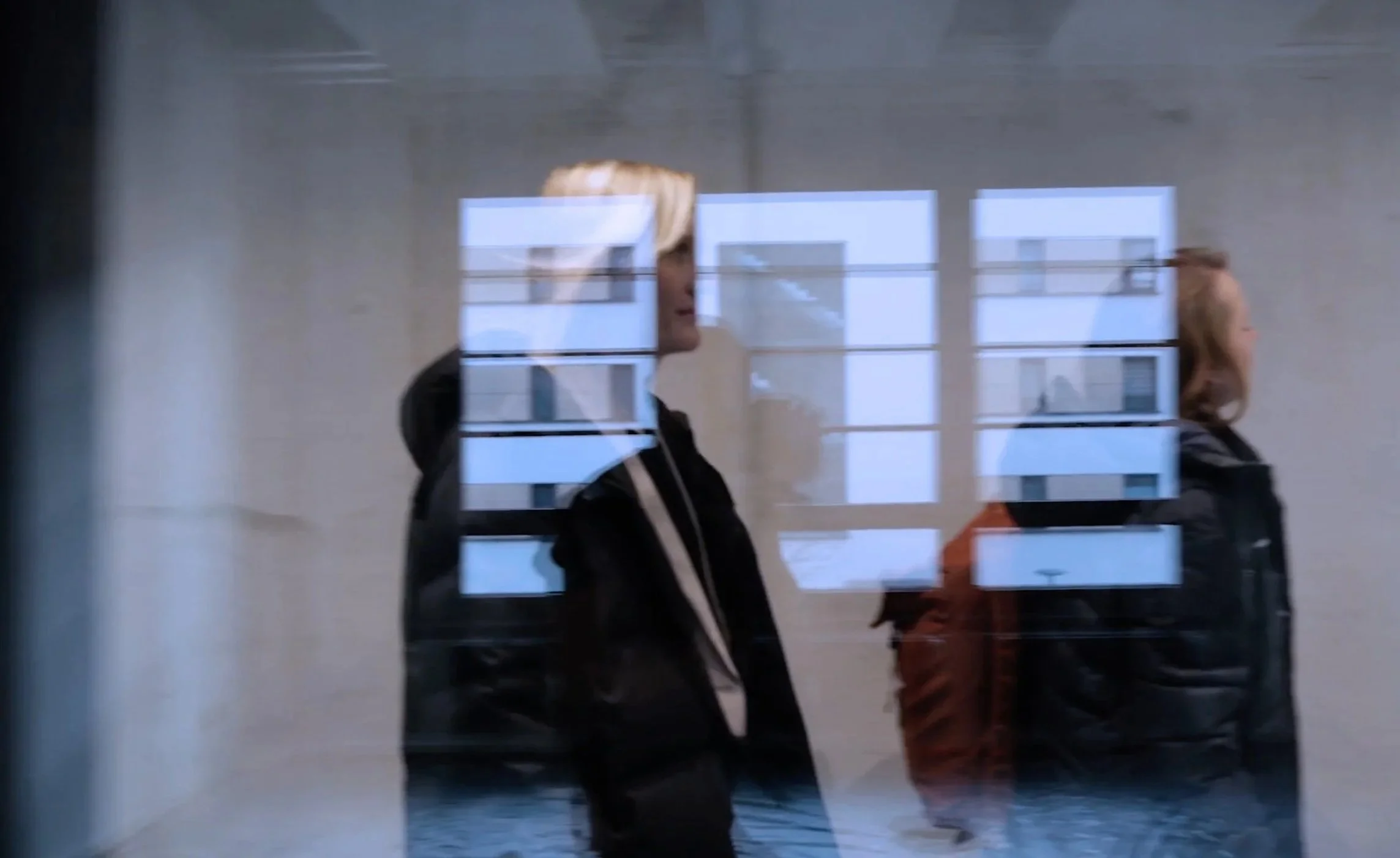 Zwei Frauen gehen an einer glasschaufensterartigen Wand mit reflektierenden Fenstern vorbei.