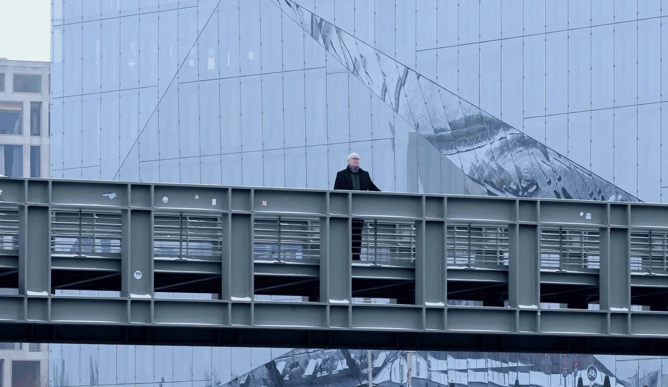 Ein Mann steht auf einer Fußgängerbrücke vor einer modernen Glasfassade mit reflektierendem Design.