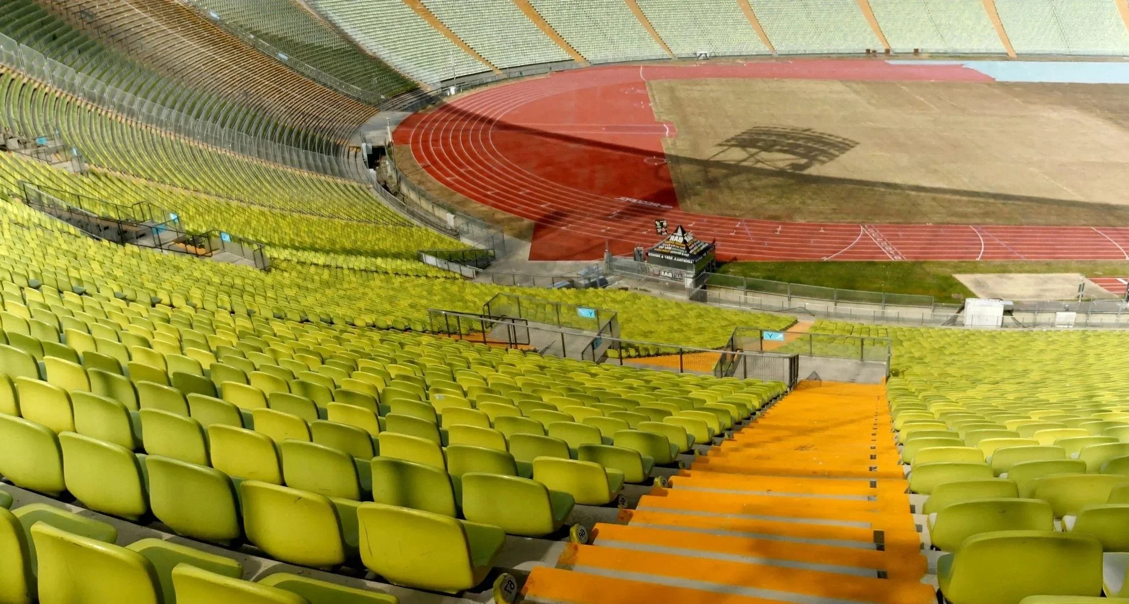 Leere grüne Zuschauerplätze in einem Stadion mit rotbraunem Laufbahn und Spielfeld, bei Sonnenlicht, Blick von oben nach unten.
