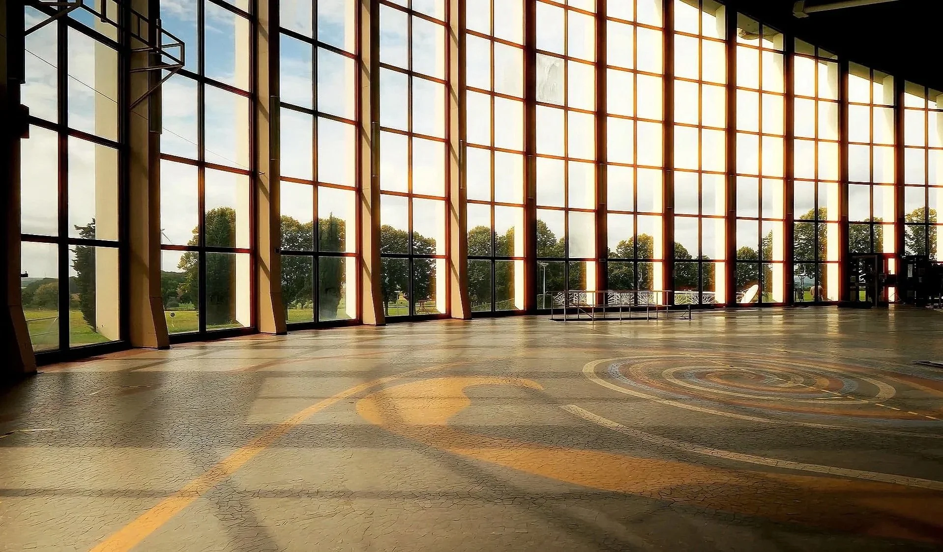 Großer, heller Raum mit hohen Fenstern, die einen Panoramablick auf eine grüne Landschaft zeigen, und einem farbigen, geometrischen Bodenmuster.