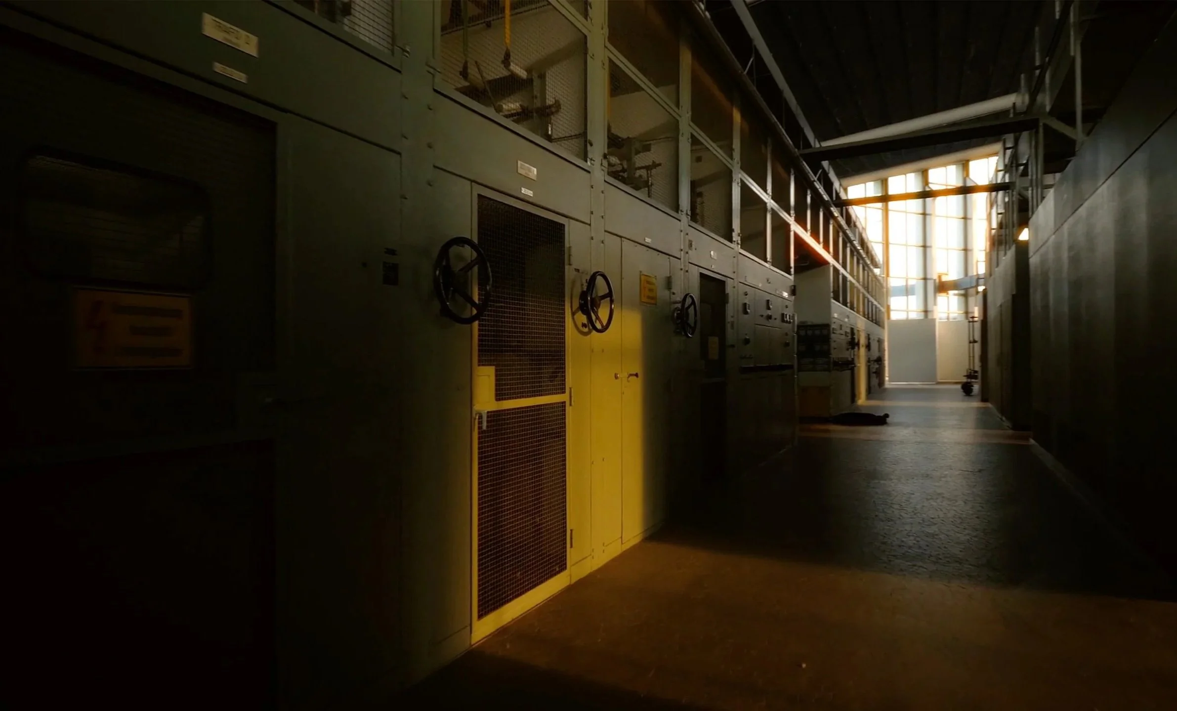 Ein dunkler Raum mit alten technischen Geräten und Steuerungskästen, durch große Fenster im Hintergrund scheint Licht herein.