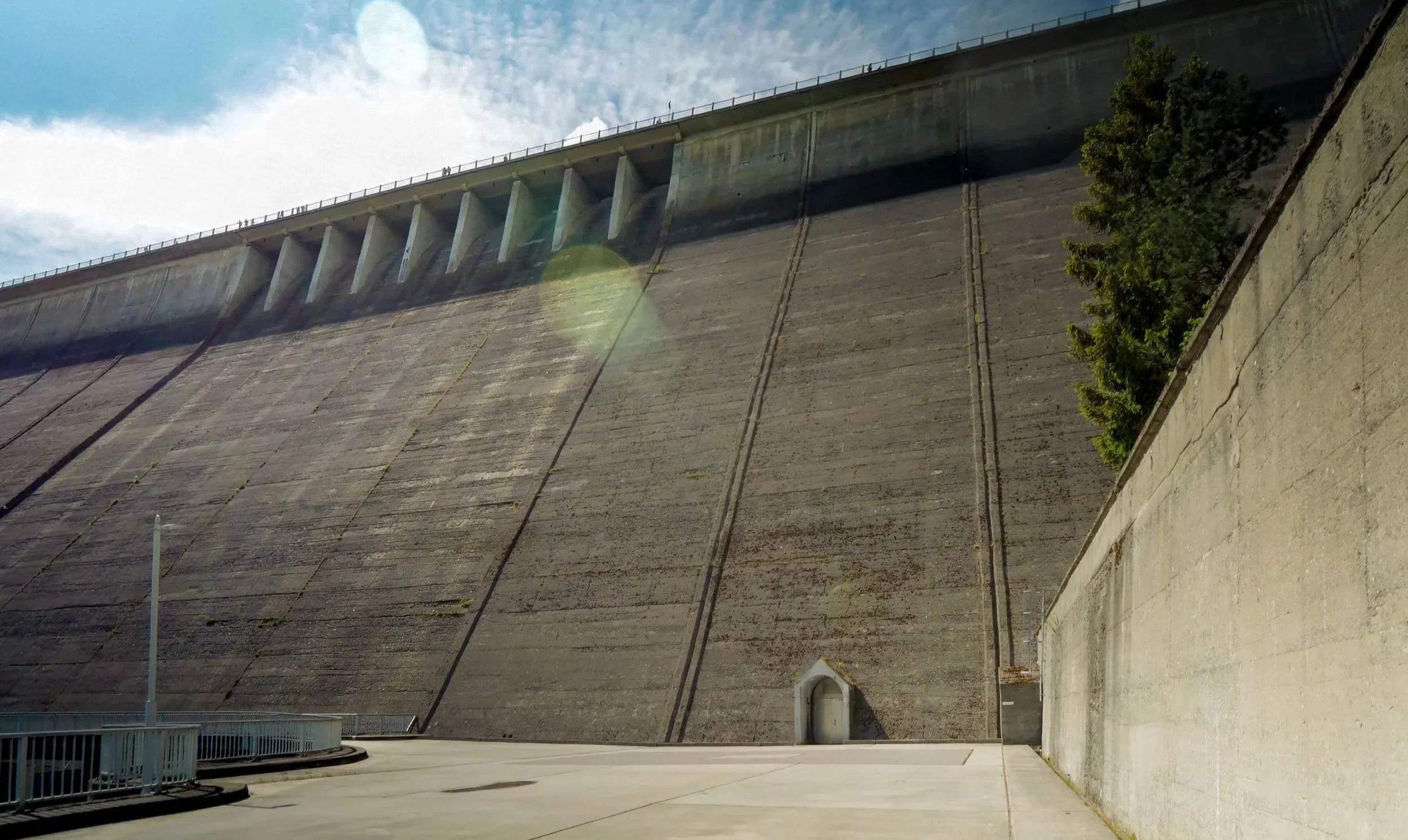 Ein großer Staudamm aus Beton mit Wasserabflussrinnen auf der Oberseite und eine kleine Durchfahrt am unteren Ende, umgeben von einer Straße und einem Zaun, mit einem Baum auf der rechten Seite.