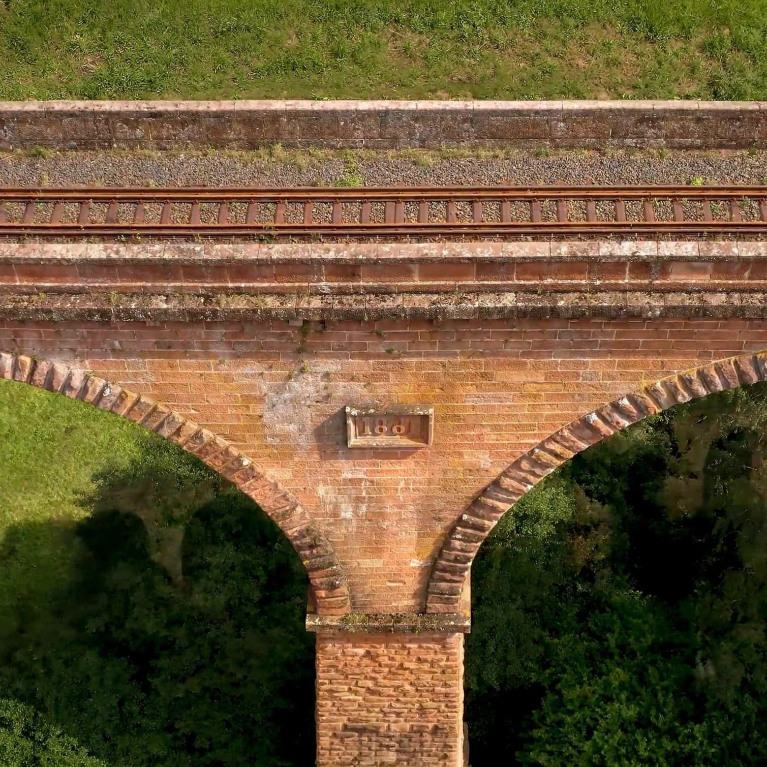 Blick auf einen alten roten Brückenbogen mit Eisenbahnstrecke darüber, umgeben von grünen Bäumen und Wiesen.