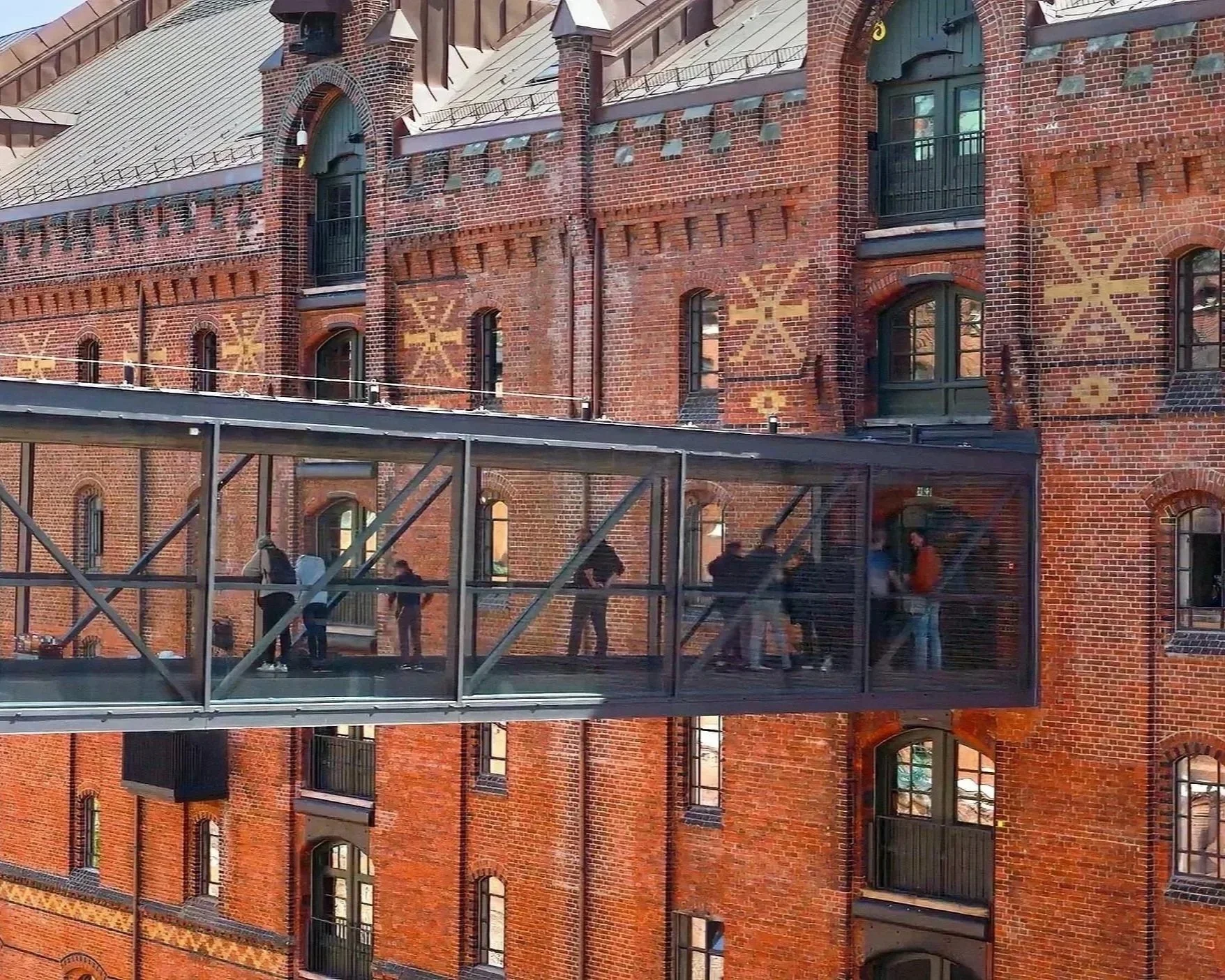 Menschen gehen über eine gläserne Brücke vor einem roten Backsteingebäude mit decorativen Mustern an der Fassade.