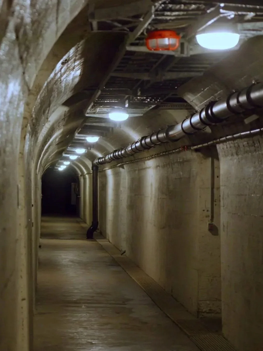 Ein dunkler, langer Tunnel mit Beleuchtung an der Decke, Wasserleitung und Gitter am Boden.