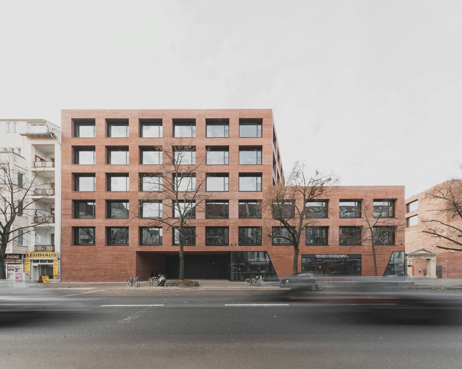 Modernes rotes Backstein-Gebäude mit großen rechteckigen Fenstern, Bäumen im Vordergrund, parkenden Fahrrädern und Autos, an einer Straße in einer Stadt