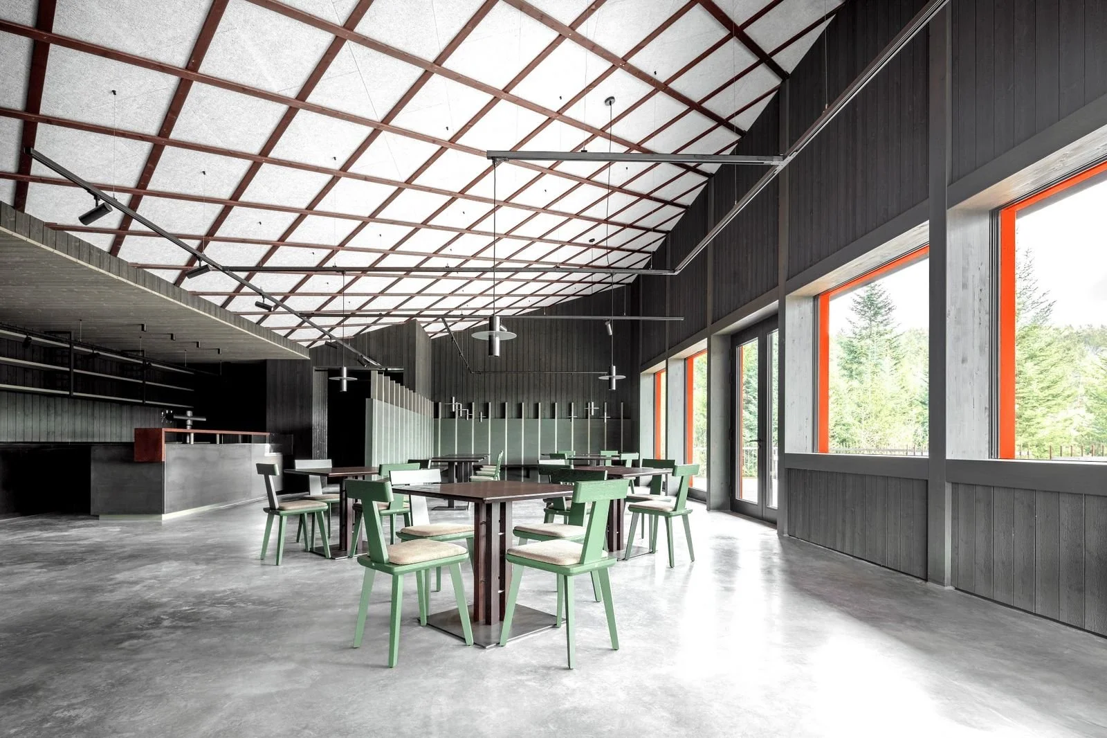 Ein leerer Restaurant- oder Cafébereich mit Holztischen, grünen Stühlen, großen Fenstern mit orange Rahmen und einer hohen Decke mit Holz- und Metallstrukturen.