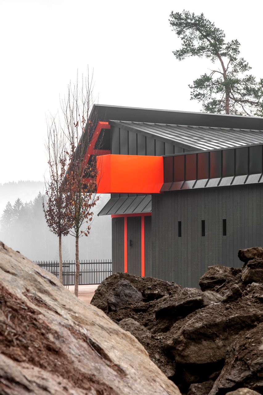 Ein modernes, schwarzes Haus mit orangefarbenen Akzenten und einem Metalldach, umgeben von Felsen und Bäumen in nebliger Landschaft.