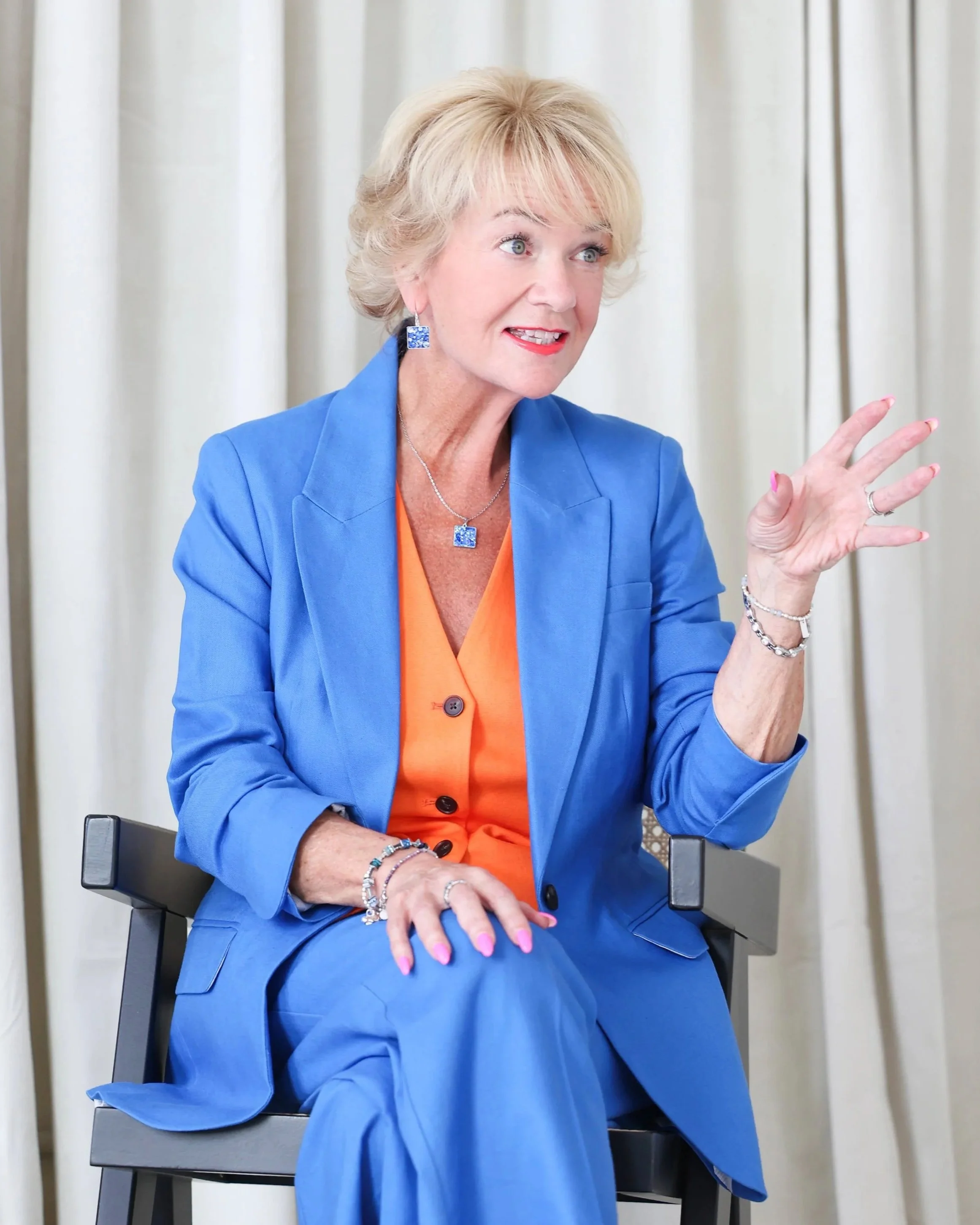Jane sitting in a chair, animatedly talking to someone. Wearing an electric blue suit and burnt orange waistcoat