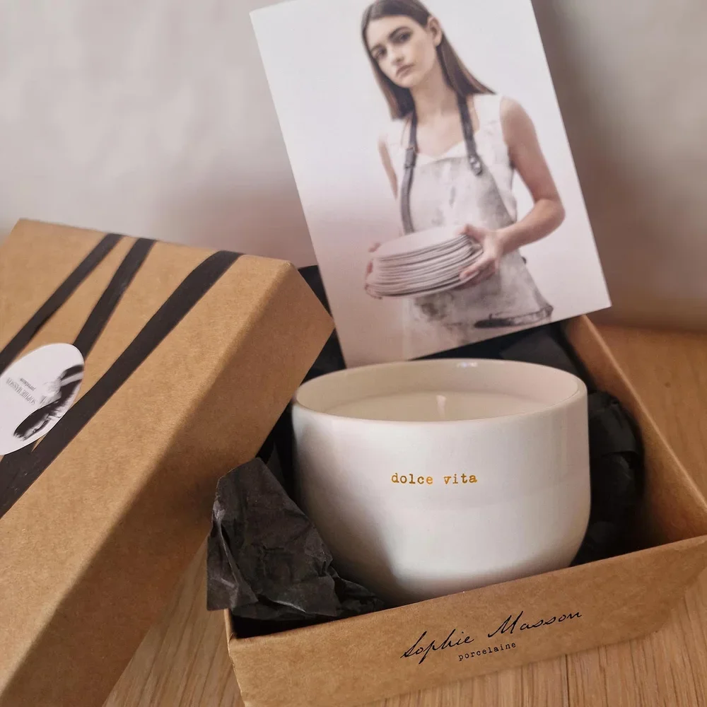 Une boîte en carton contenant une tasse en porcelaine blanche avec l'inscription 'dolce vita', un morceau de papier noir, et une photo d'une femme portant un tablier, tenant un plateau de plusieurs assiettes.
