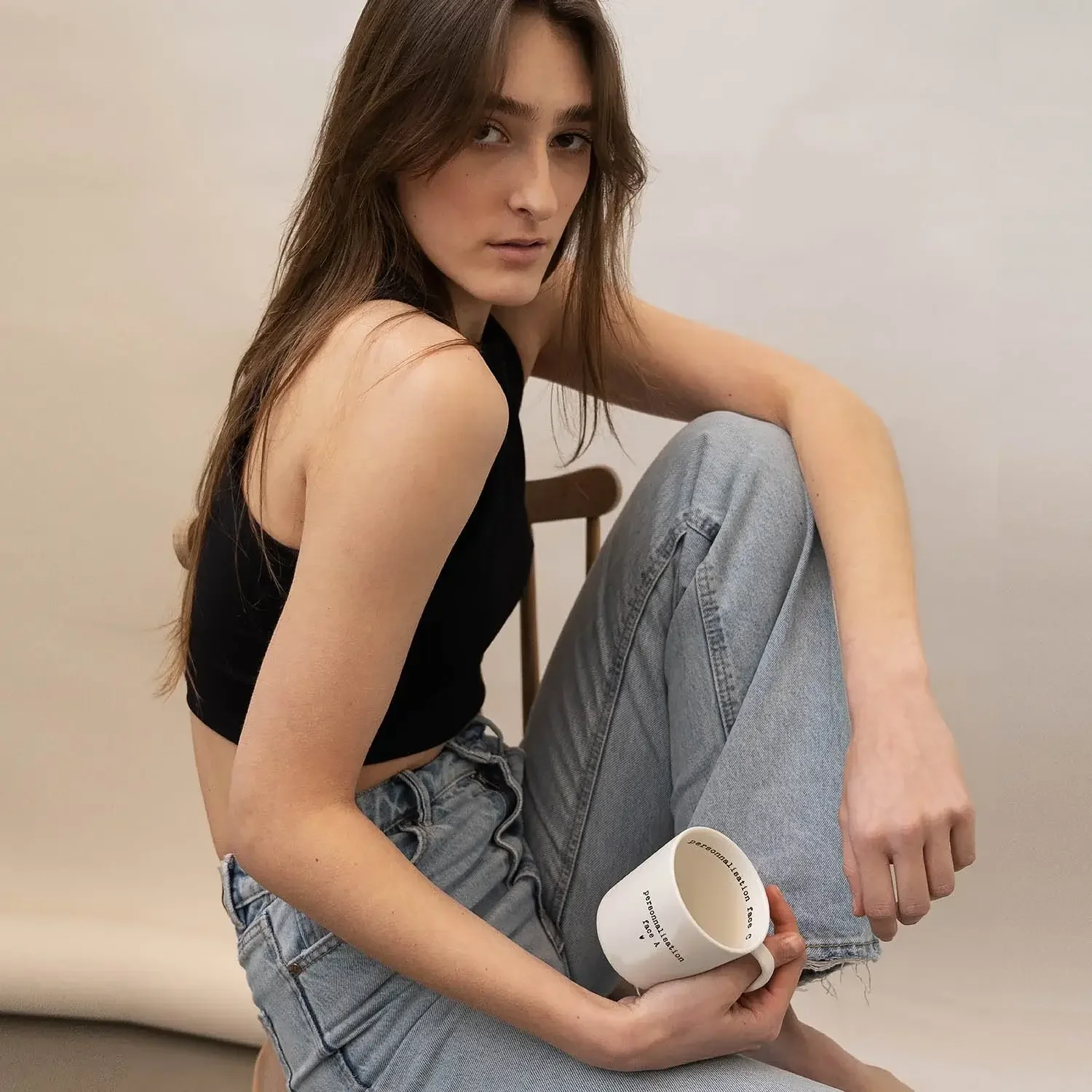 Femme aux cheveux bruns, portant un haut noir et un jeans déchiré, assise contre un mur beige, tenant une tasse blanche dans sa main.