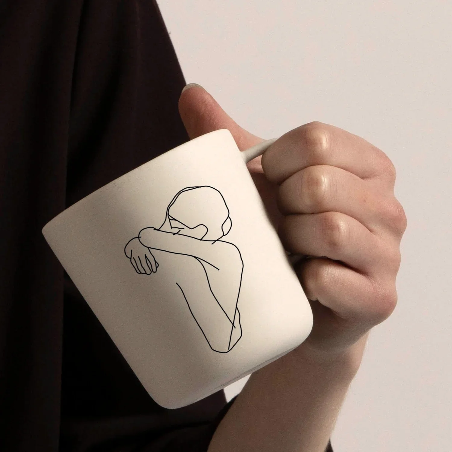 Une main tient une tasse blanche avec une illustration d'une personne en train d'embrasser ou de pleurer avec des bras croisés, sur un fond blanc.