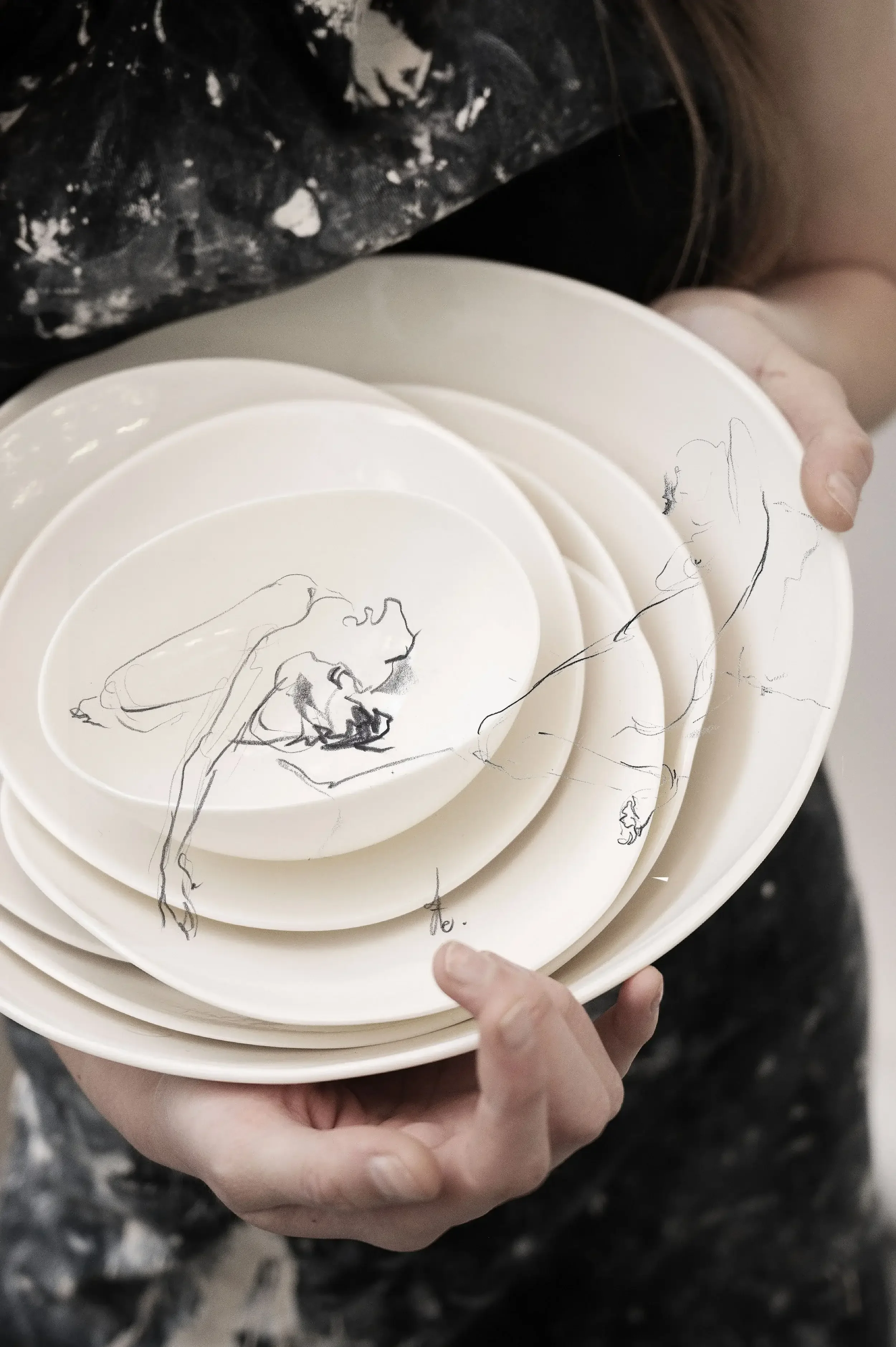 Une personne tient plusieurs assiettes blanches empilées, avec un dessin d'une femme assise qui semble danser ou se pencher. La personne porte un vêtement avec un motif marbré noir et blanc.