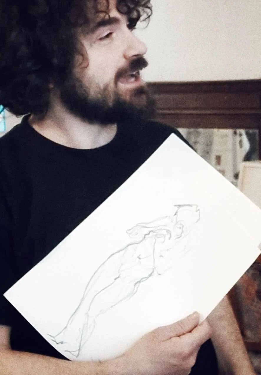 Un homme avec des cheveux bouclés et une barbe tient un dessin de femme nue. La scène se déroule probablement dans une maison ou un restaurant.