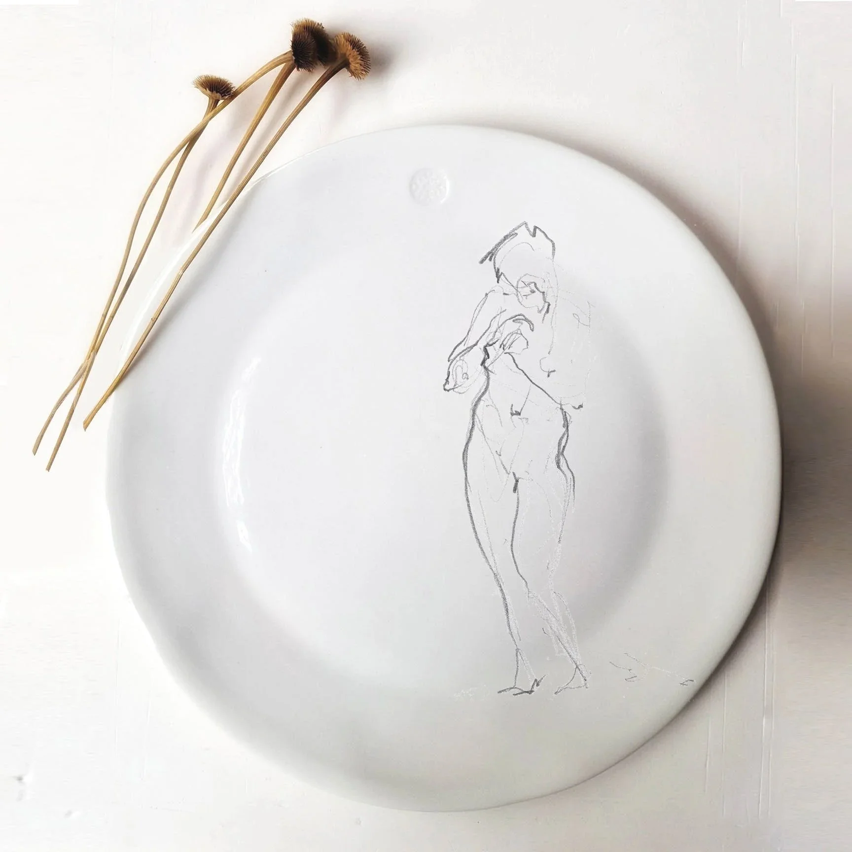 Assiette blanche avec un dessin de femme en ligne simple et quelques tiges de fleurs séchées naturelles posées sur le bord.