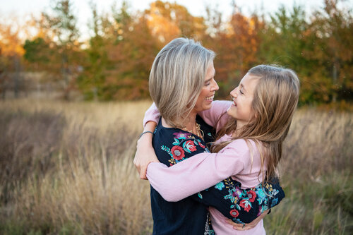A woman and girl hug in an autumn field with tall grasses and colorful trees