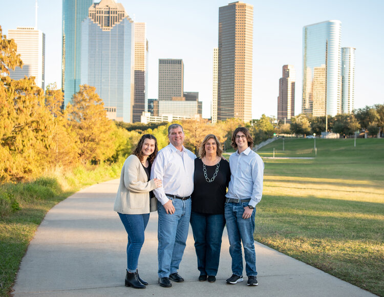 A family of four stands on a path with a city skyline and autumn trees in the background