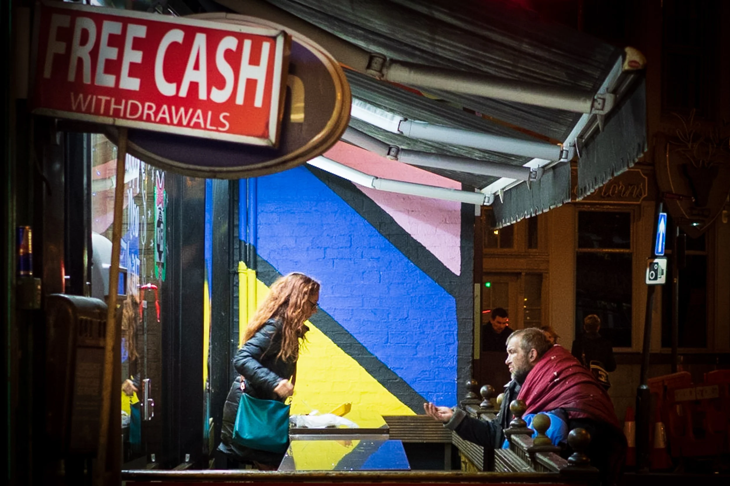 Homeless man asking for money from woman under sign saying "free cash withdrawals"