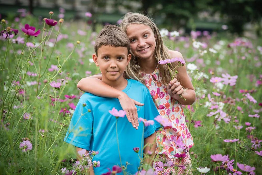 big sister has arm around brother in a field of cosmos flowers