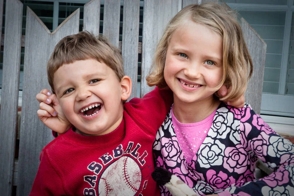 brother and sister share a laugh with their arms around each other on a bench