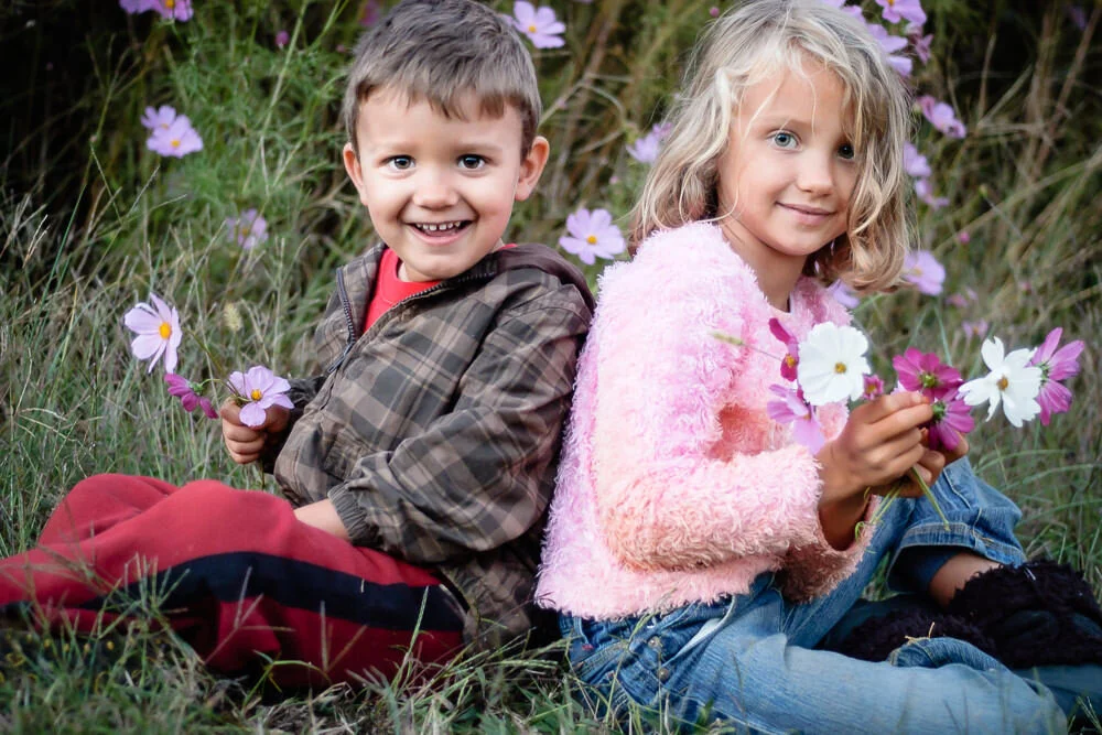 brother and sister sit back to back in a field of cosmos flowers