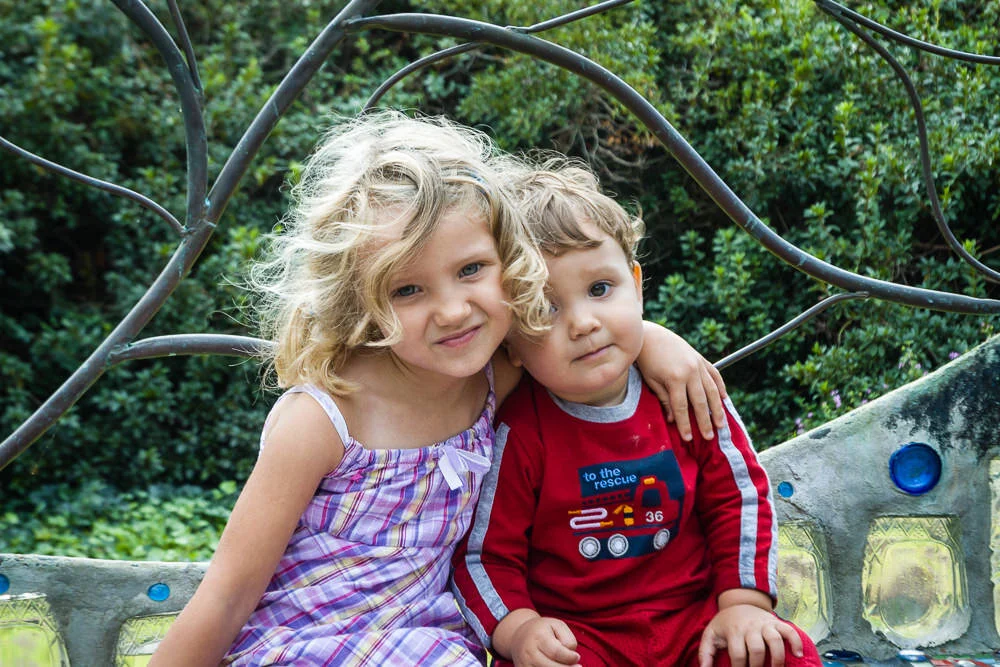 big sister has her arm around her little brother while seated on a bench