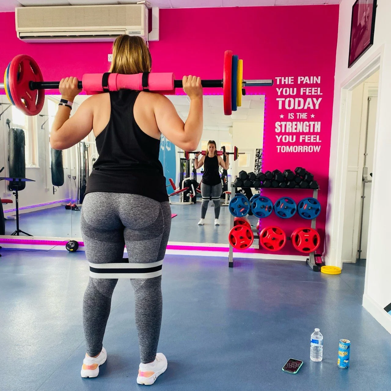 A woman lifting a barbell at a gym, with a colorful wall with weights and a motivational quote in the background.