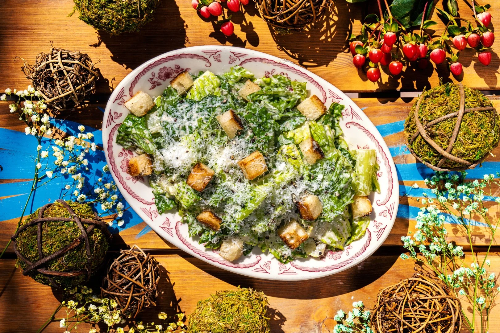 Caesar salad with croutons and parmesan on a decorative plate, surrounded by twigs, floral elements, and berries on a wooden table.