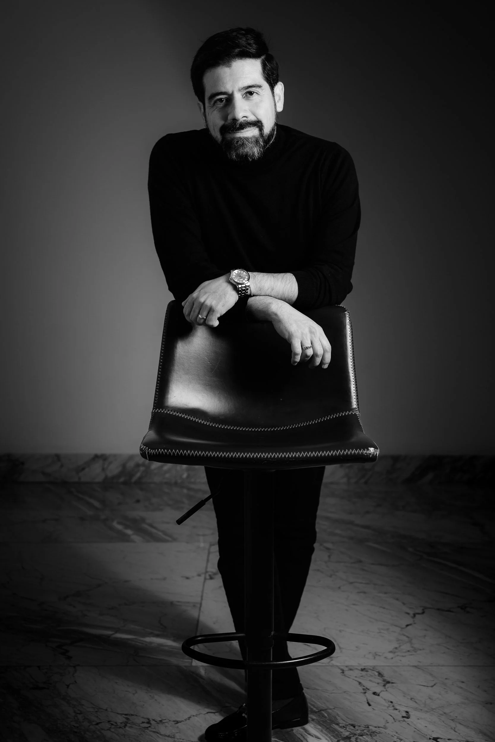 Retrato ejecutivo en blanco y negro de un hombre con suéter negro apoyado en un banco de piel, sobre piso de mármol. Fotografía por Rodrigo Ojeda.