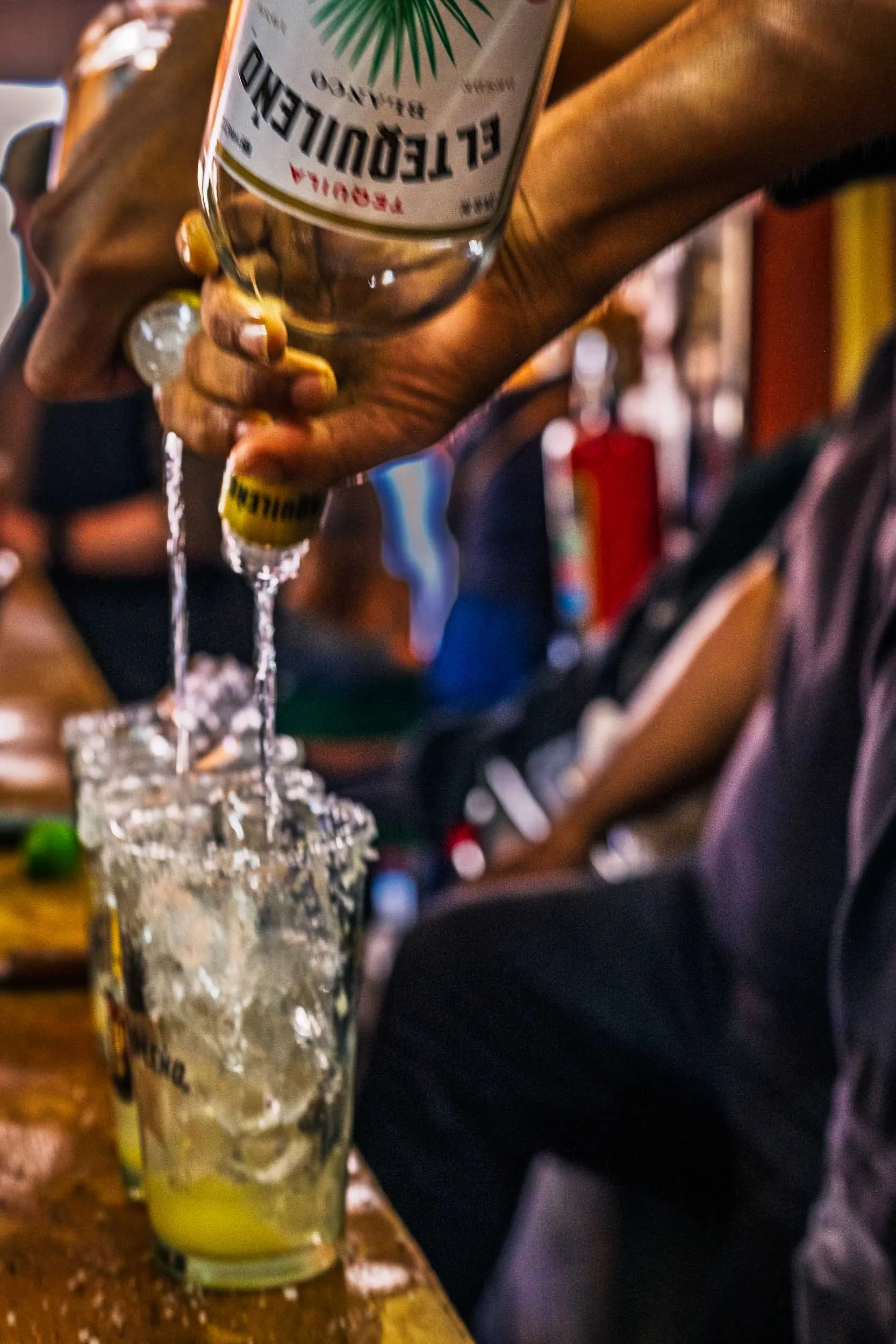 Mano vertiendo tequila El Tequileño en vasos con hielo en un bar tradicional, escena para reportaje cultural o editorial gastronómica.