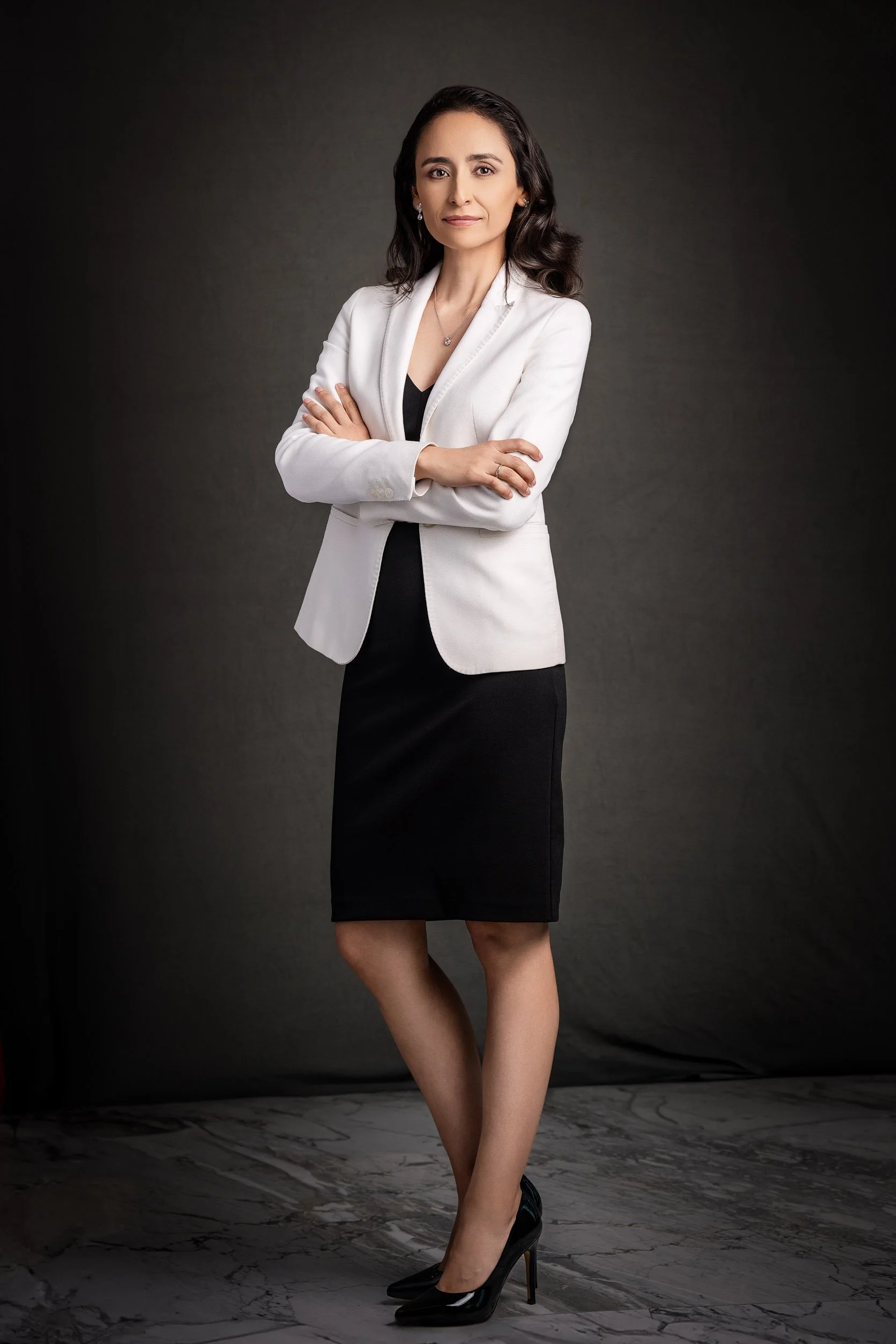 Retrato ejecutivo de cuerpo completo de una mujer con blazer blanco y vestido negro, brazos cruzados, en estudio con fondo oscuro. Fotografía por Rodrigo Ojeda.