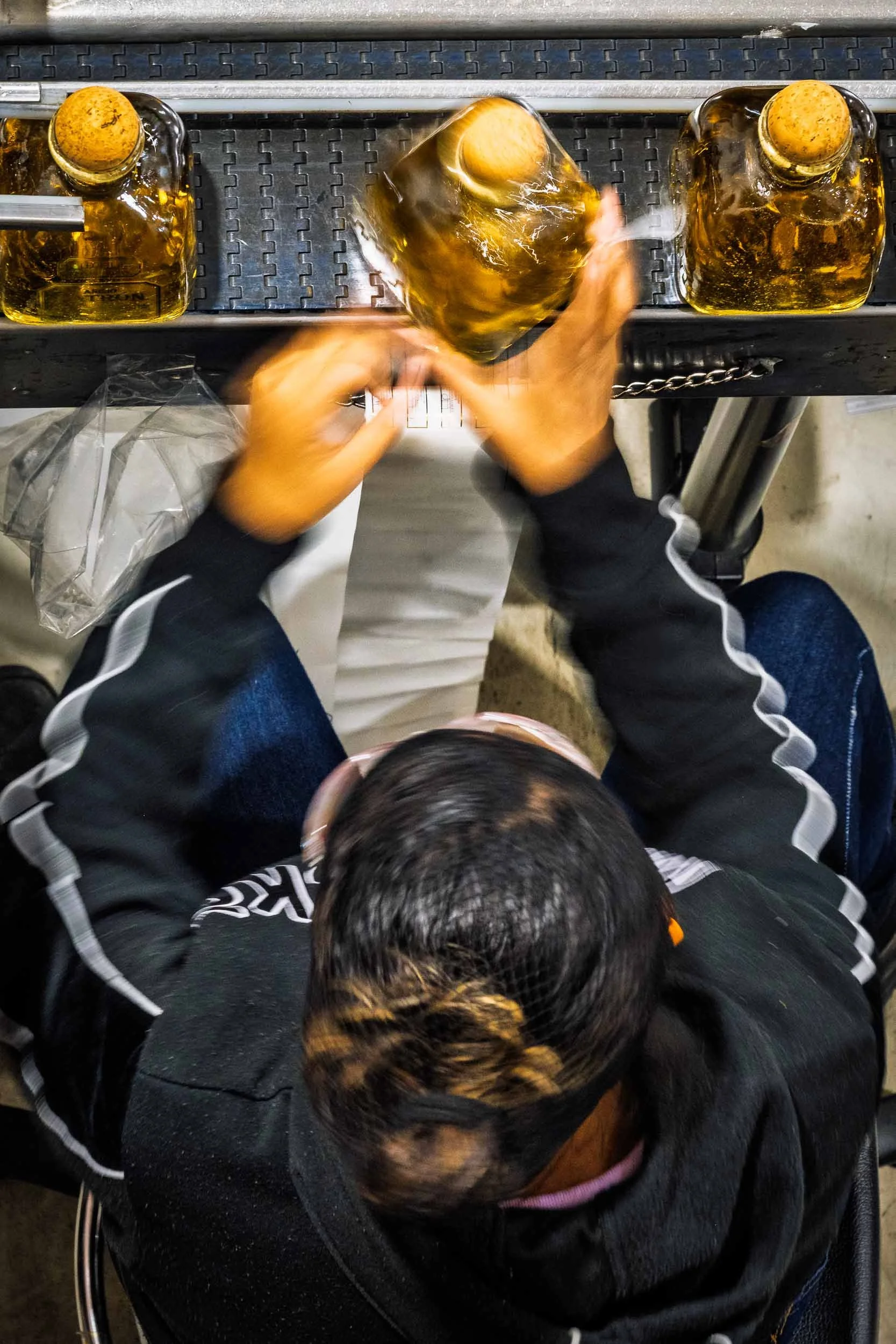 Vista cenital de trabajadora etiquetando botellas de tequila a mano en una fábrica, imagen representativa de proceso artesanal.