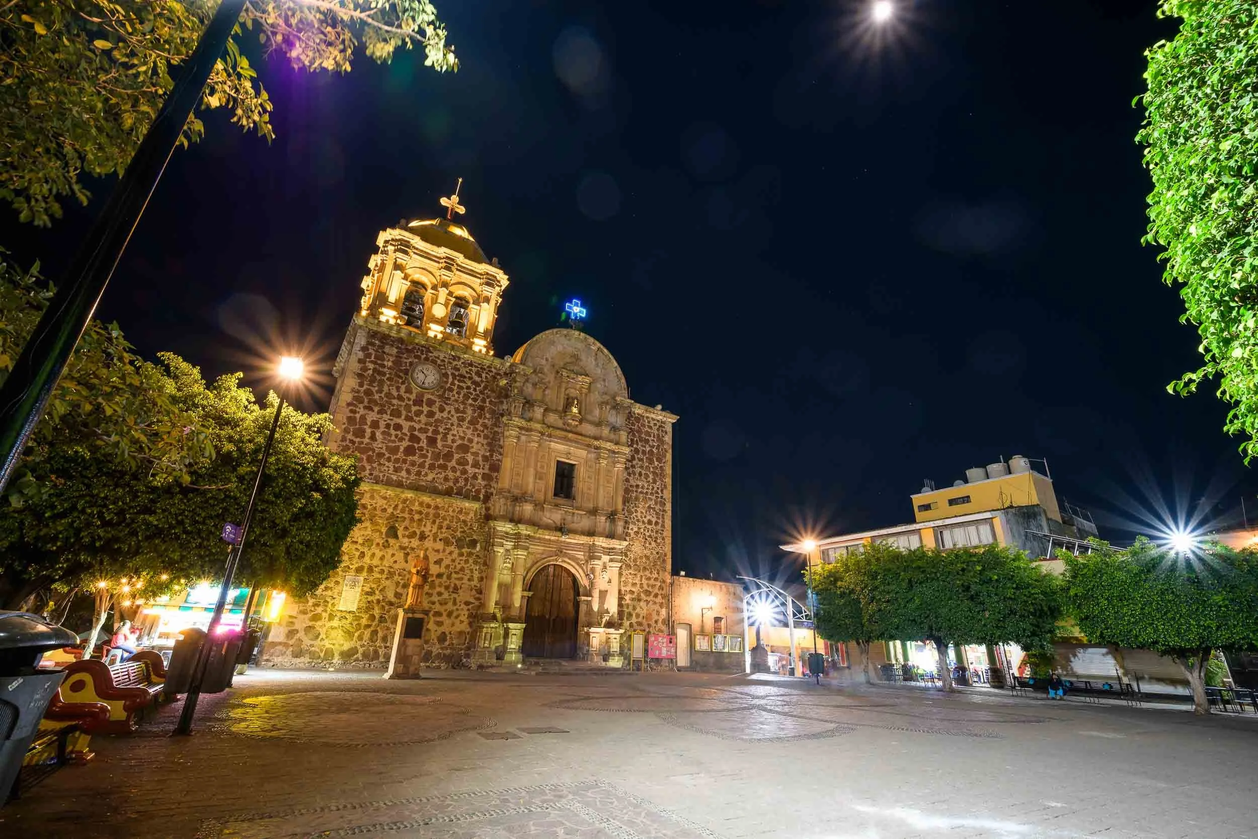 Vista panorámica del pueblo de Tequila con arquitectura colonial iluminada de noche, incluyendo una iglesia principal y árboles frondosos en la plaza central.