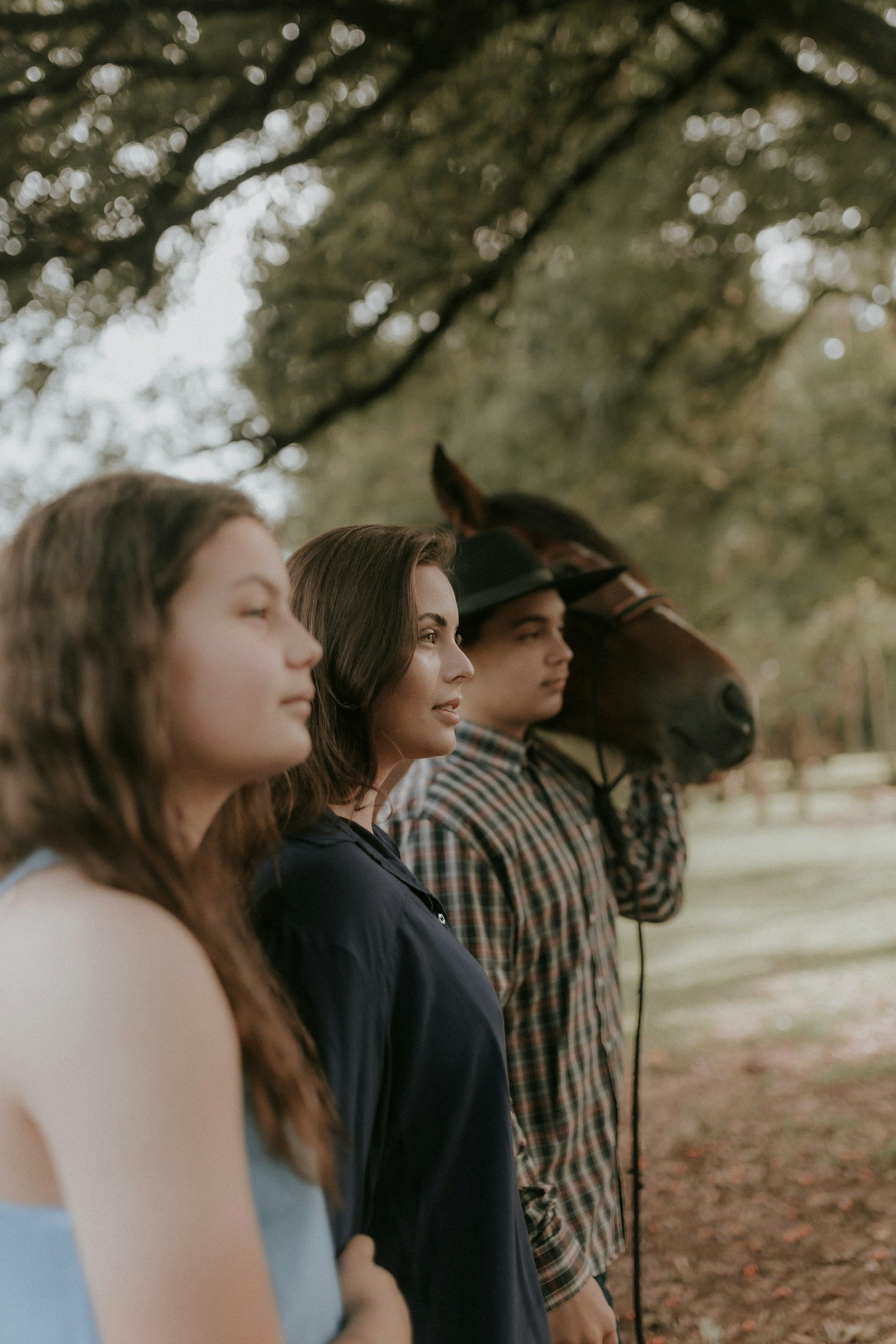 A line of young people standing outdoors in a park or wooded area, with trees overhead, sitting patiently and looking forward.