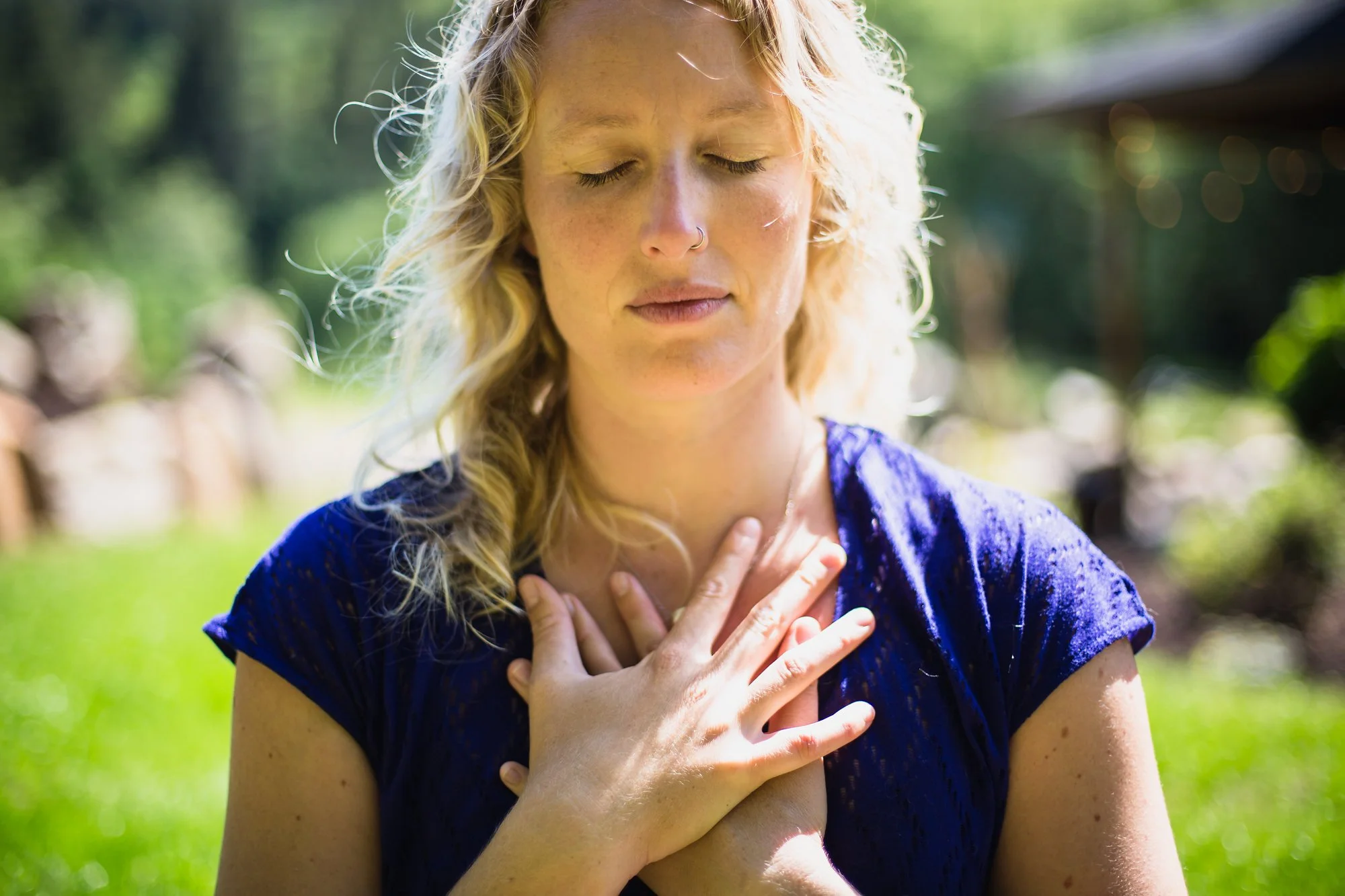 A woman with blonde curly hair, closed eyes, and a nose ring standing outdoors with her hands crossed over her chest, wearing a blue dress.