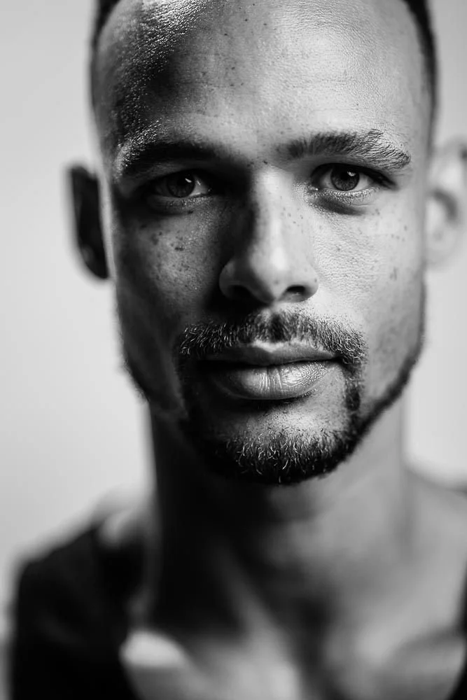 Close-up black and white portrait of a young man with short hair, beard, and light skin, looking directly into the camera.