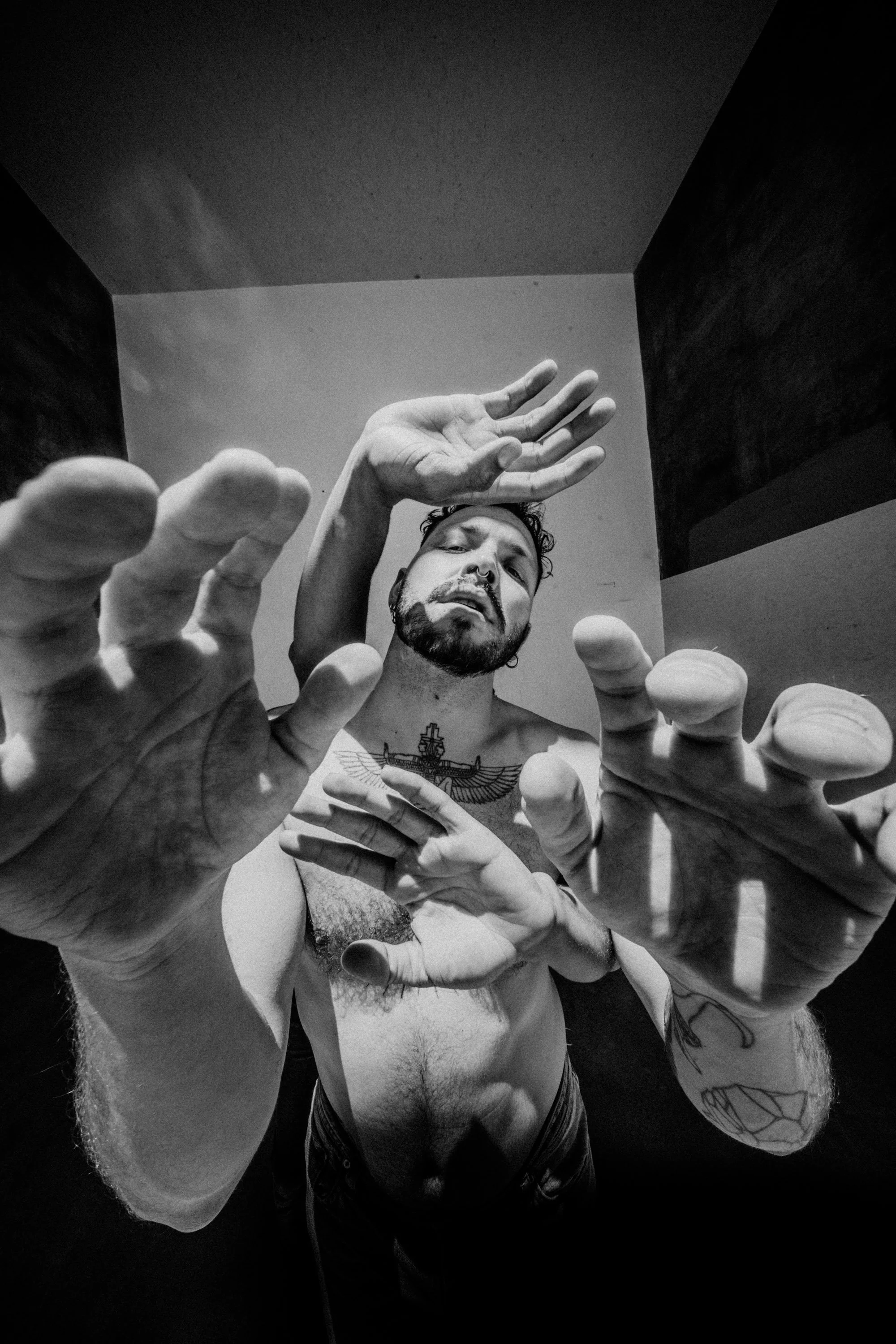 Black and white photo of a shirtless man with tattoos, outstretched hands reaching towards the camera, taken from a low angle outdoors.