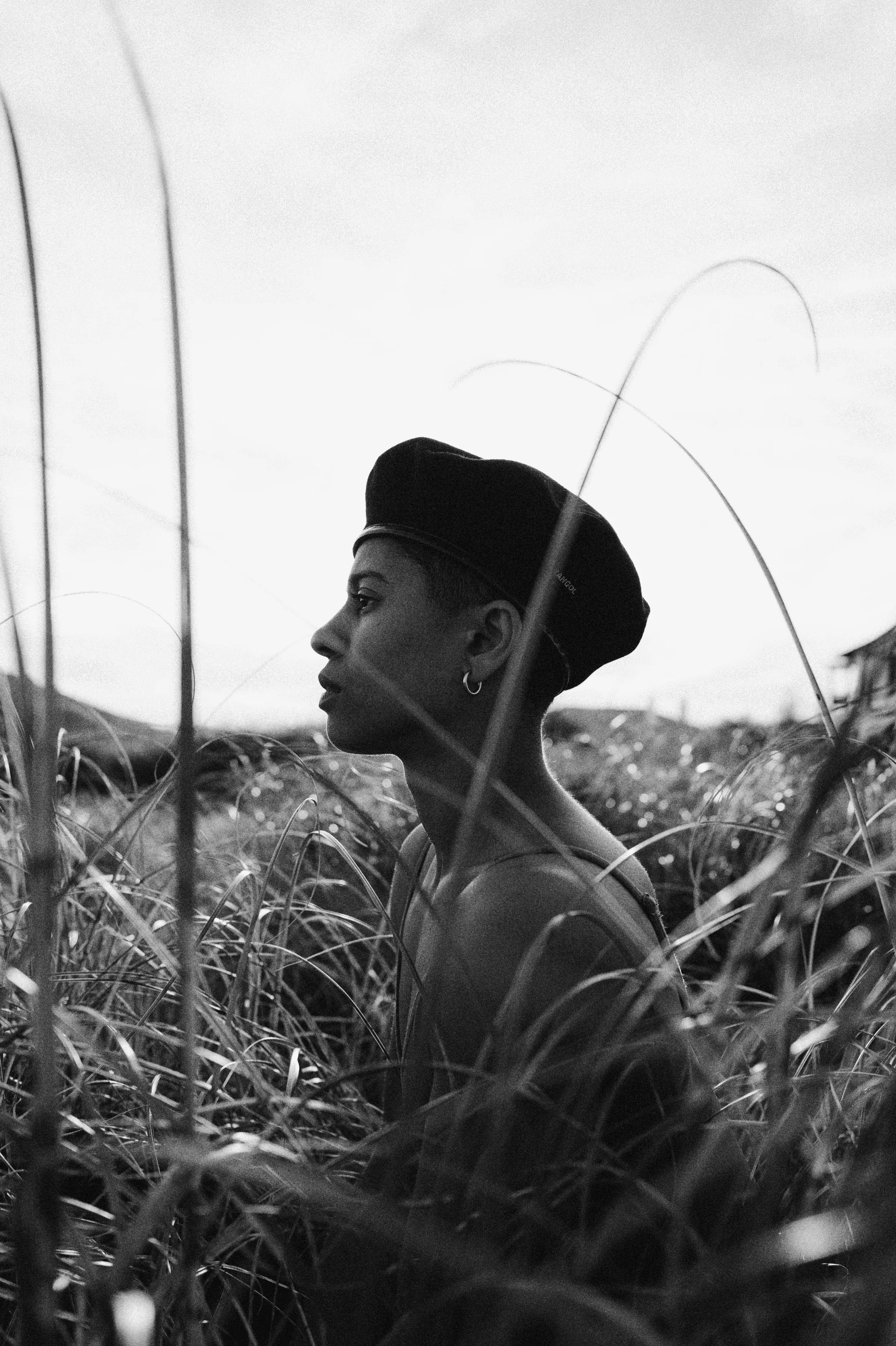 A woman with earrings and a beret standing in tall grass, captured in black and white with a cloudy sky background.