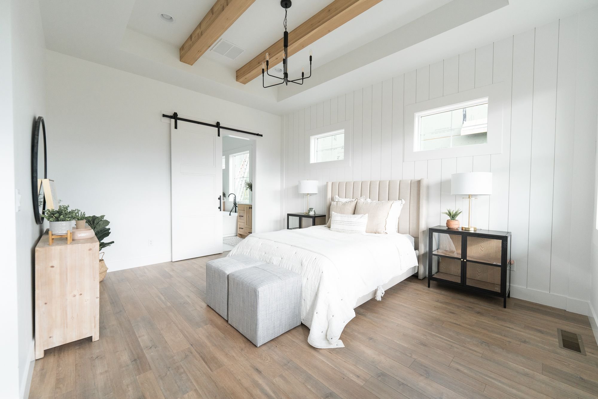 Modern bedroom with light wooden flooring, featuring a bed with white bedding, two gray ottomans at the foot, and black side tables with lamps. A sliding door leads to a bathroom. White walls, wooden ceiling beams, and decorative plants complete the