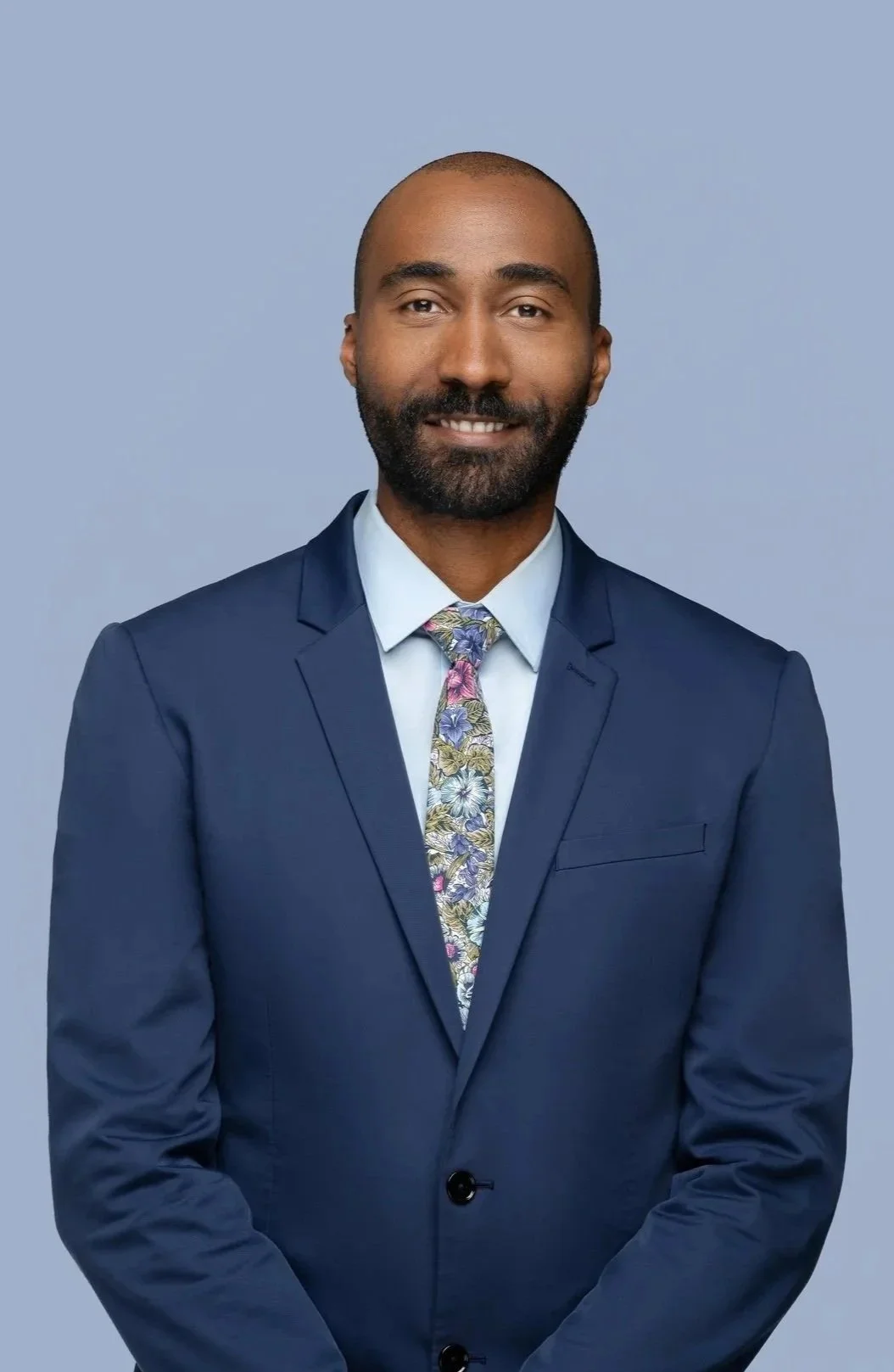 A man in a navy blue suit, light blue shirt, and a colorful floral tie, smiling against a plain grey background.