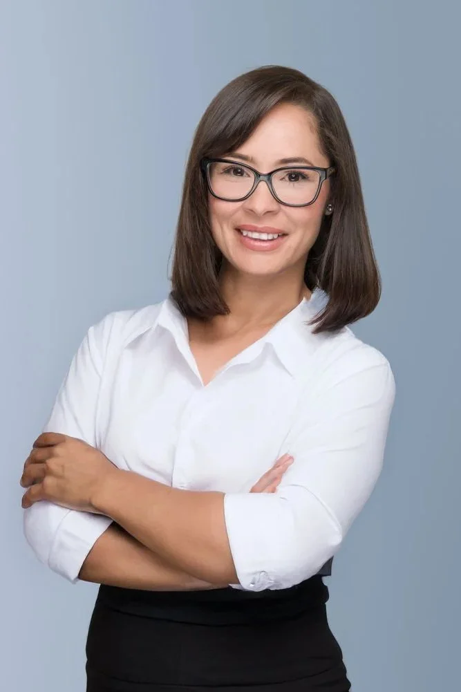 A woman with shoulder-length dark hair, wearing glasses, a white shirt with lace details on the collar and cuffs, and smiling with arms crossed, standing against a light blue background.