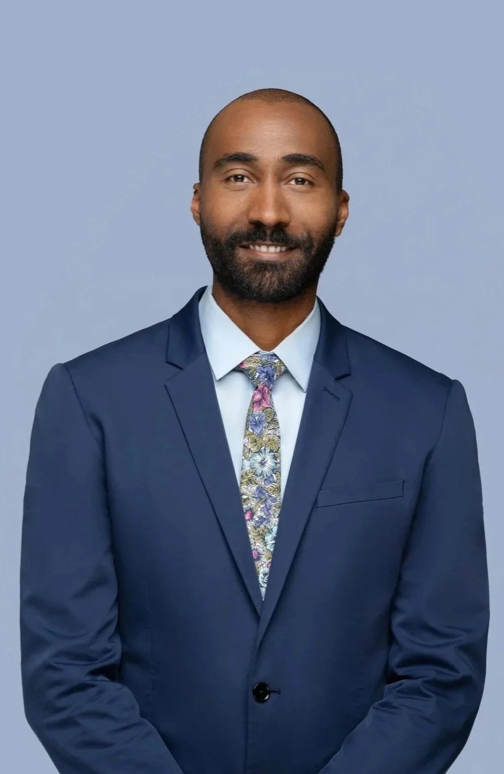 A man in a navy blue suit, light blue shirt, and a colorful floral tie, smiling against a plain grey background.