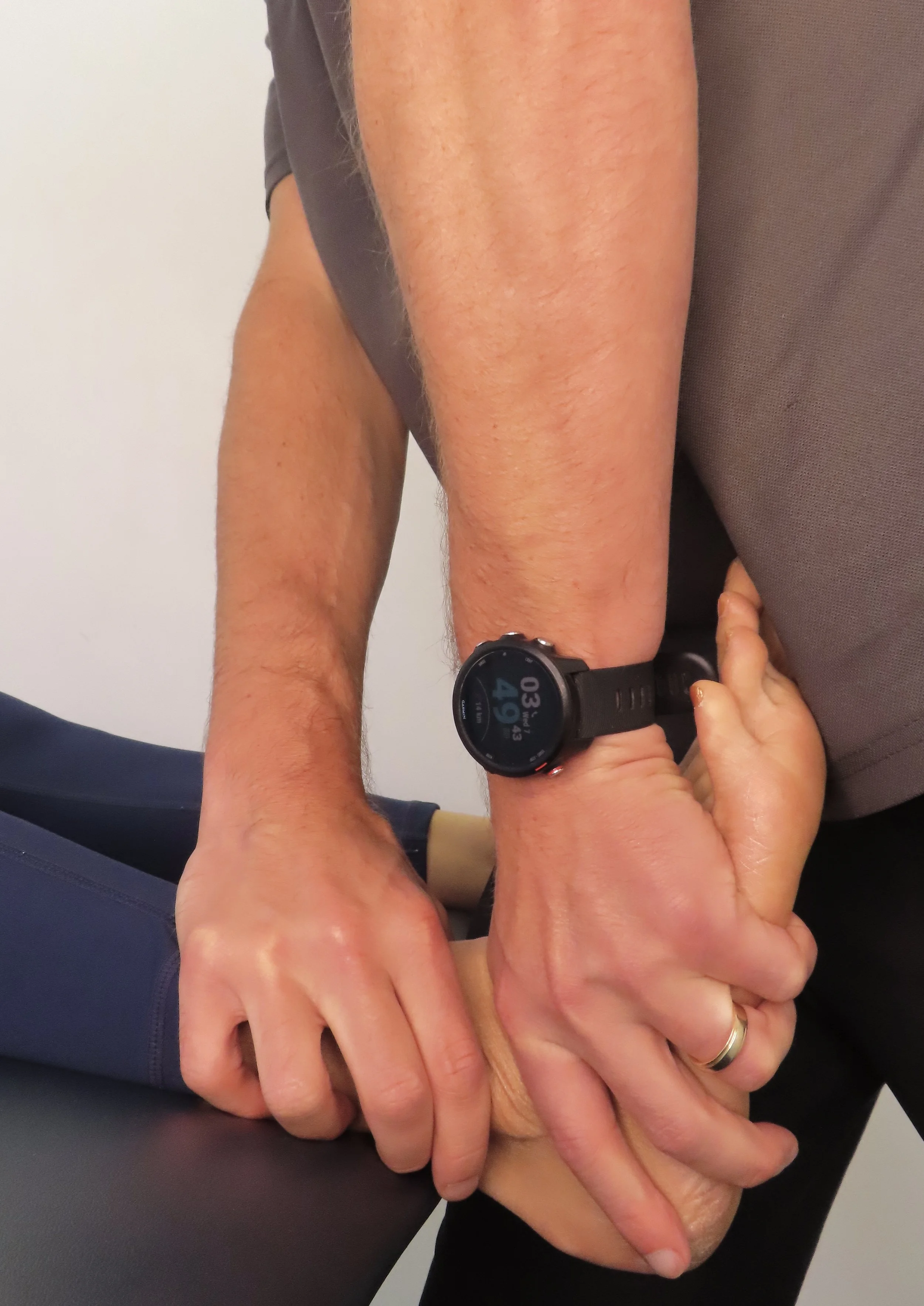 An individual is receiving a physical therapy or medical examination, with a healthcare professional holding their hand and wrist, while wearing a smartwatch showing the time 03:49, and the patient is in a seated or reclined position.
