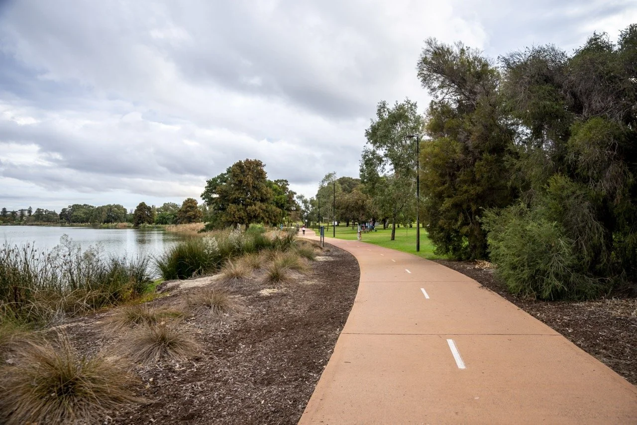 Local people walking along a shared walking and cycling path beside native bush and wetlands at Lake Galup near Mount Hawthorn, WA, under overcast skies