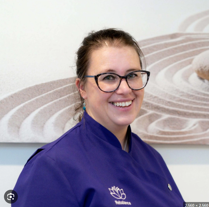 A woman with glasses, brown hair, and earrings smiling, wearing a purple uniform with a lotus flower logo and standing in front of a sandy zen garden-themed wall art.