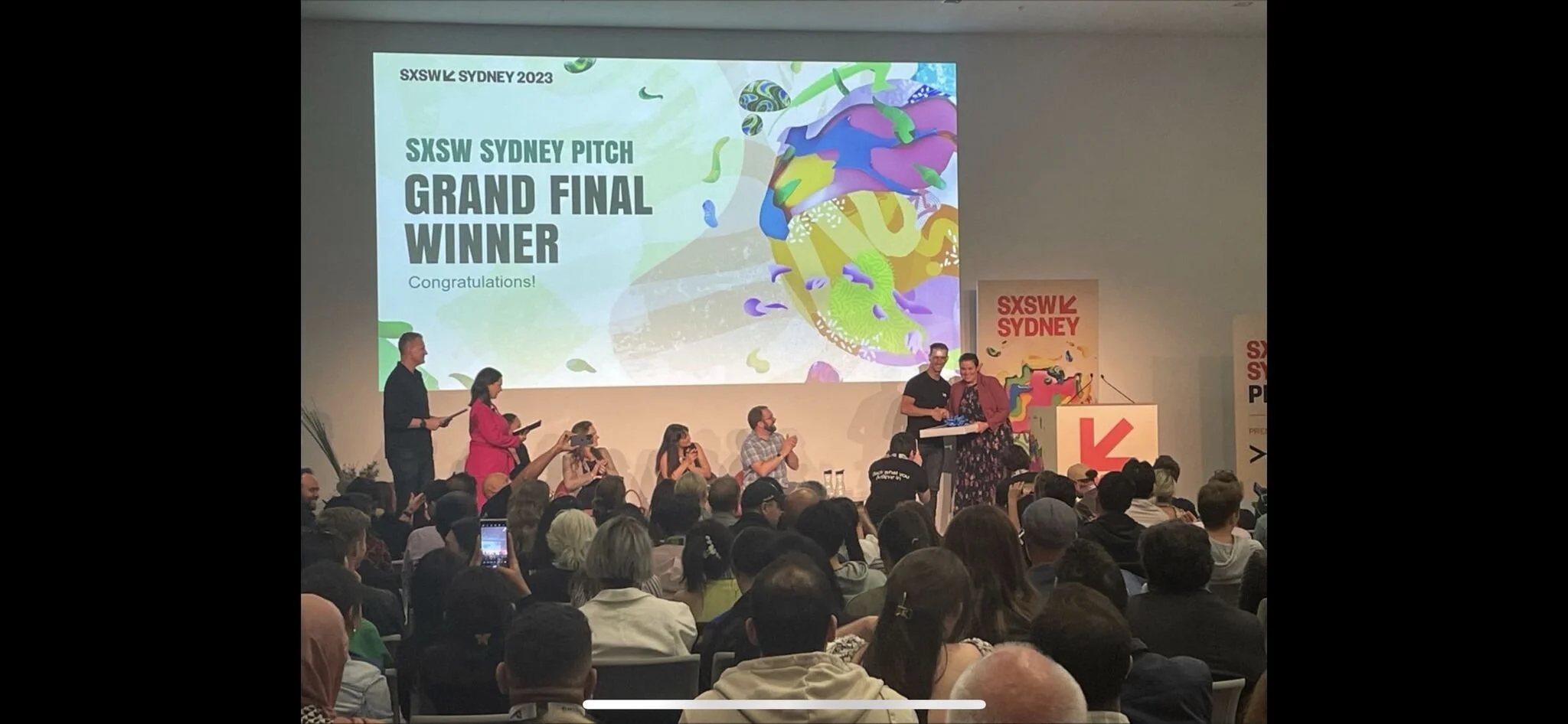 An awards ceremony at SXSW Sydney 2023, with a large crowd watching people on stage receive a prize. The screen displays a colorful graphic and the text 'SXSW Sydney Pitch Grand Final Winner. Congratulations!'