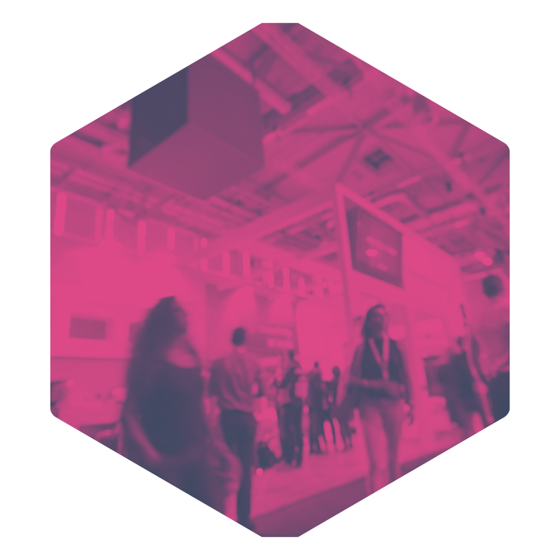 People walking in an indoor space, possibly a convention or exhibition hall with screens and overhead lights.