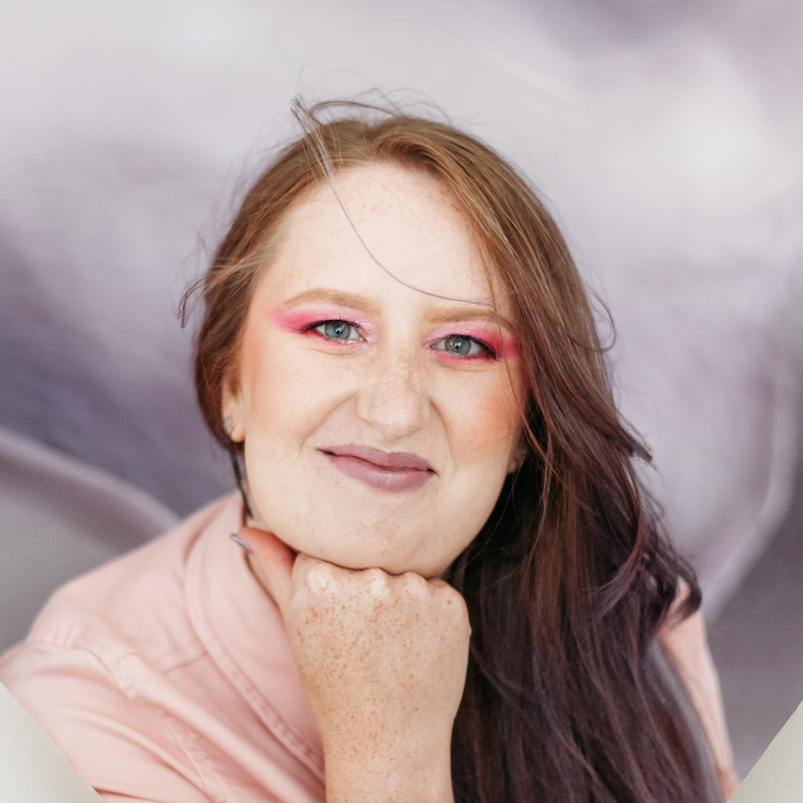 A woman with red hair, pink eyeshadow, and freckles resting her chin on her hand, smiling at the camera.