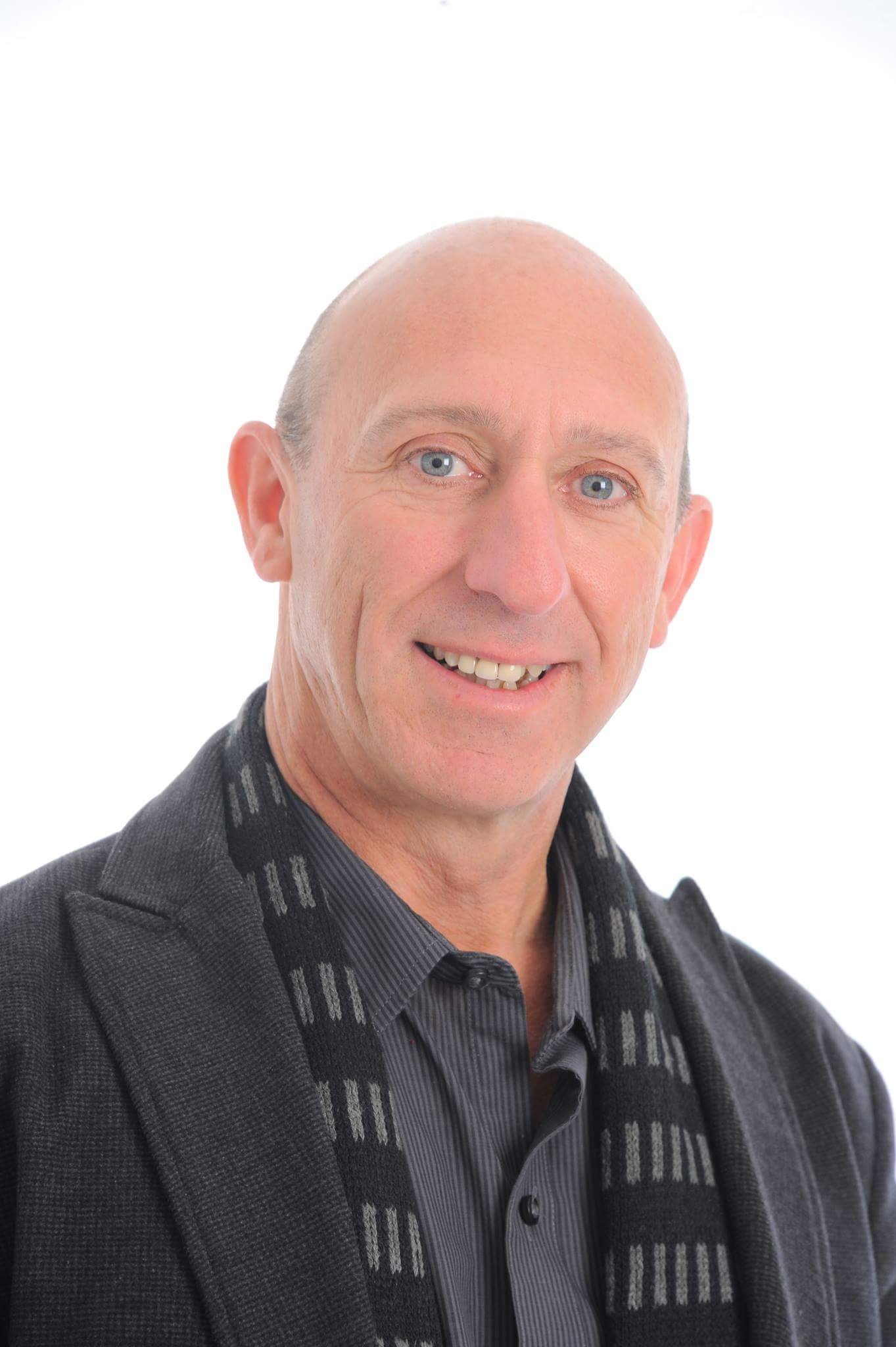 Photo of a smiling middle-aged man with a bald head, blue eyes, wearing a dark gray blazer, shirt, and scarf, against a white background.